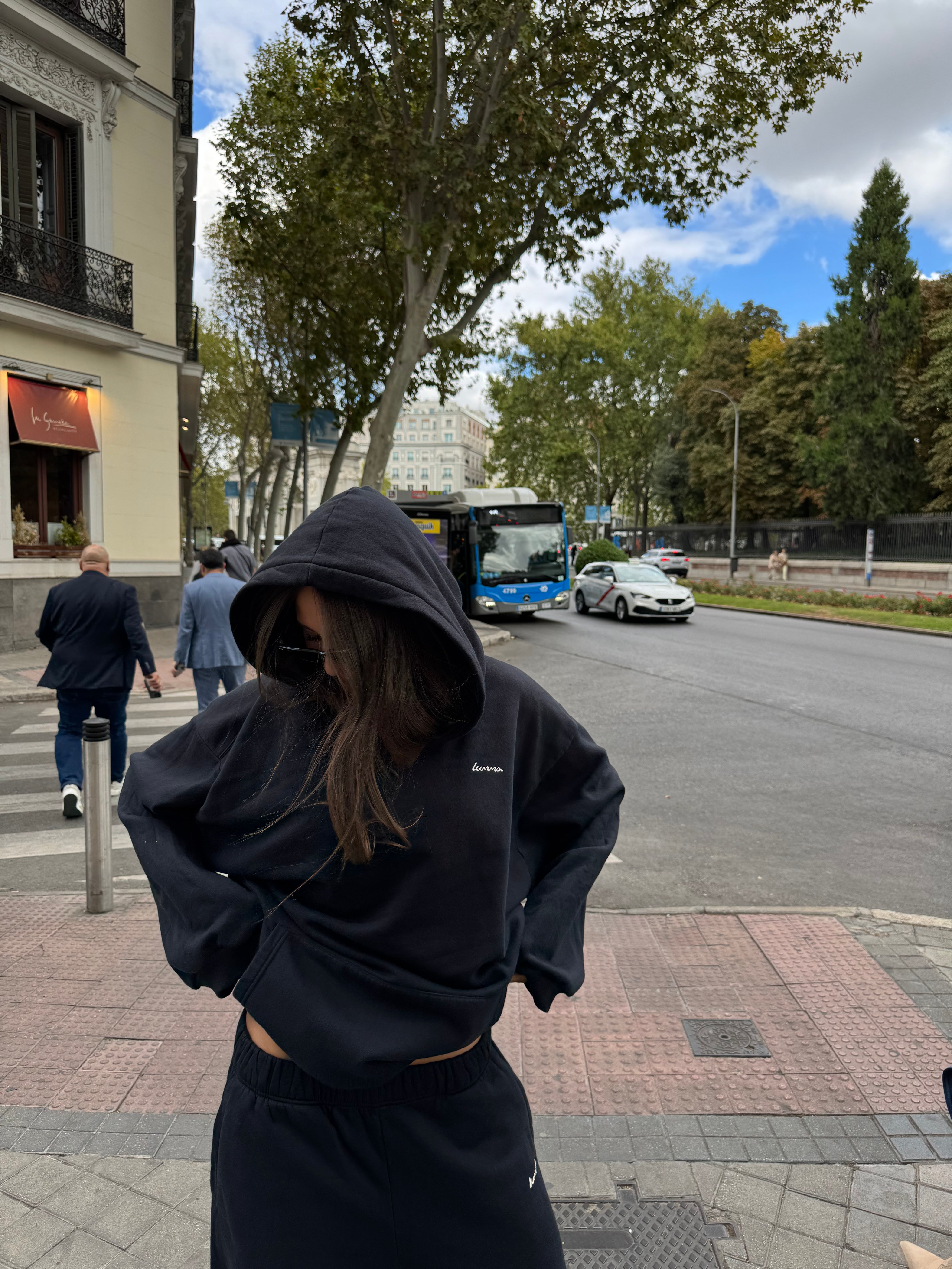 Person wearing a black hoodie on a street with trees and buildings in the background