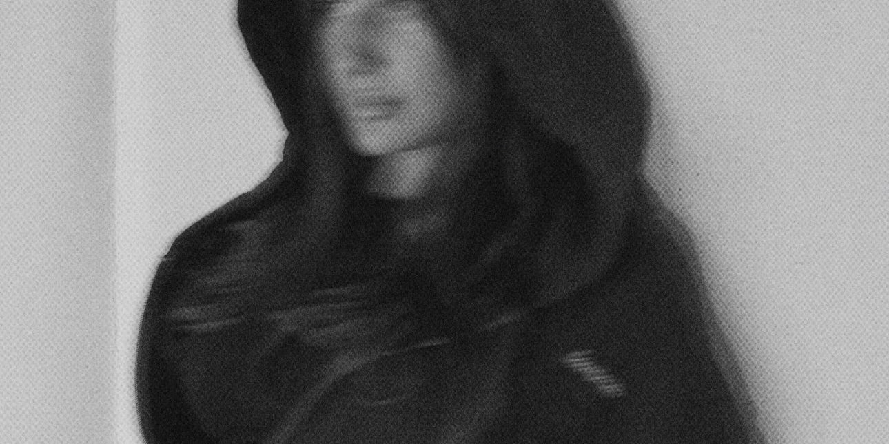 Person wearing a dark hooded garment against a plain background