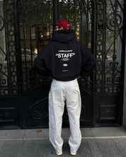Person wearing a black hoodie with 'STAFF' text, white pants, and red cap in front of an ornate gate.