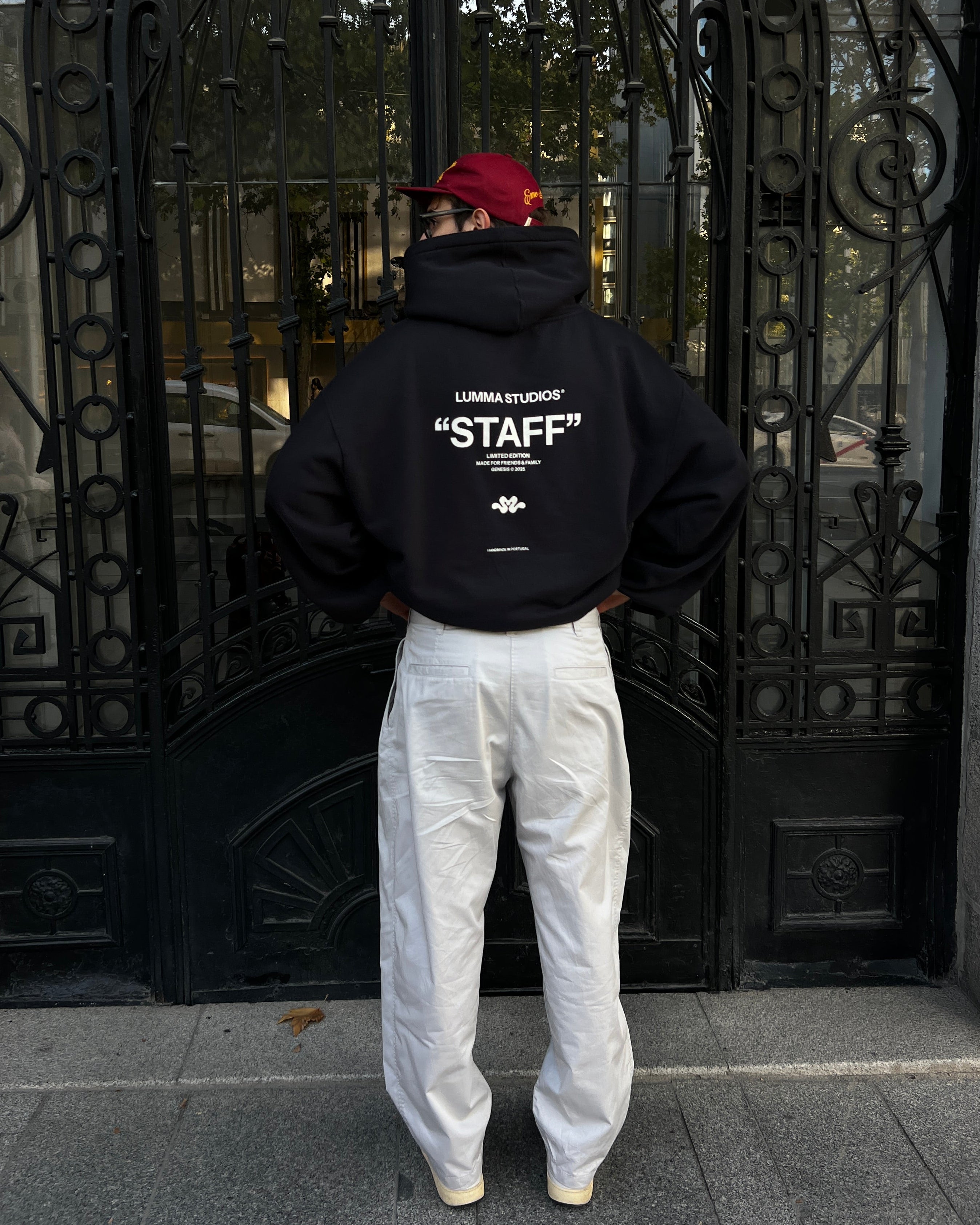 Person wearing a black hoodie with 'STAFF' text, white pants, and red cap in front of an ornate gate.