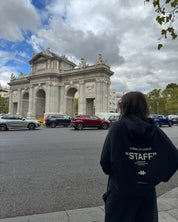 Person wearing a 'Staff' HOODIE in front of a large architectural building with a blue sky.