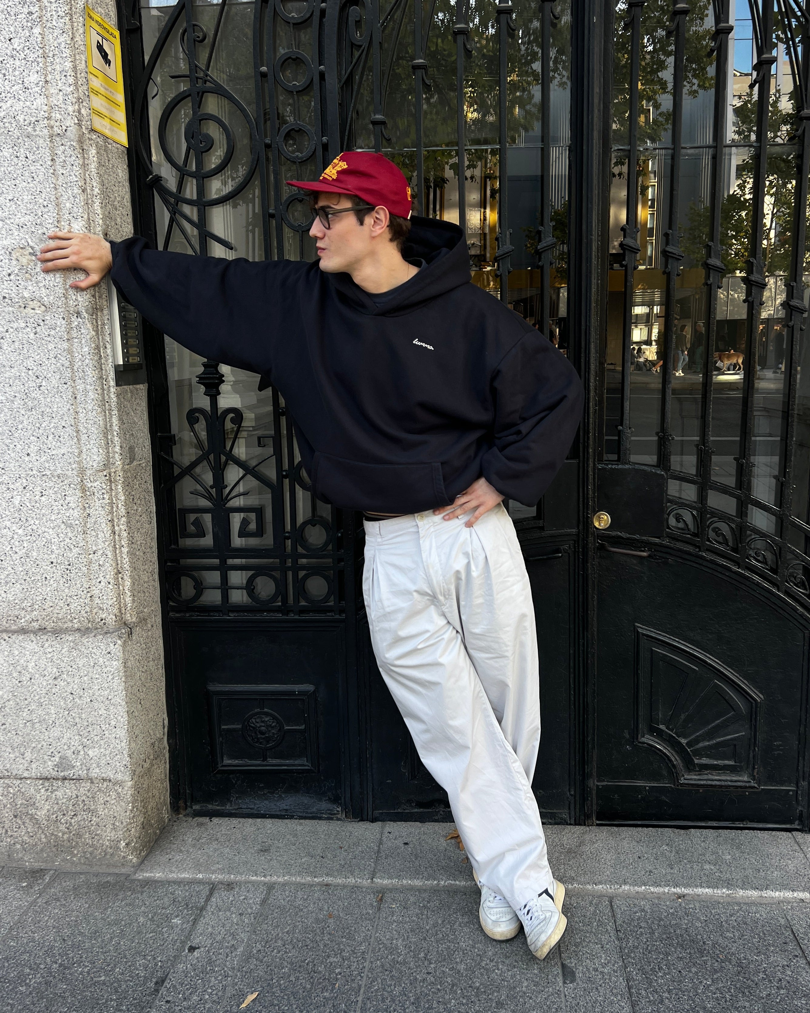 Person wearing a black hoodie, white pants, and a red cap leaning against a black metal gate.
