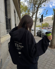 Person wearing a black hoodie with text, holding a green cup outdoors.