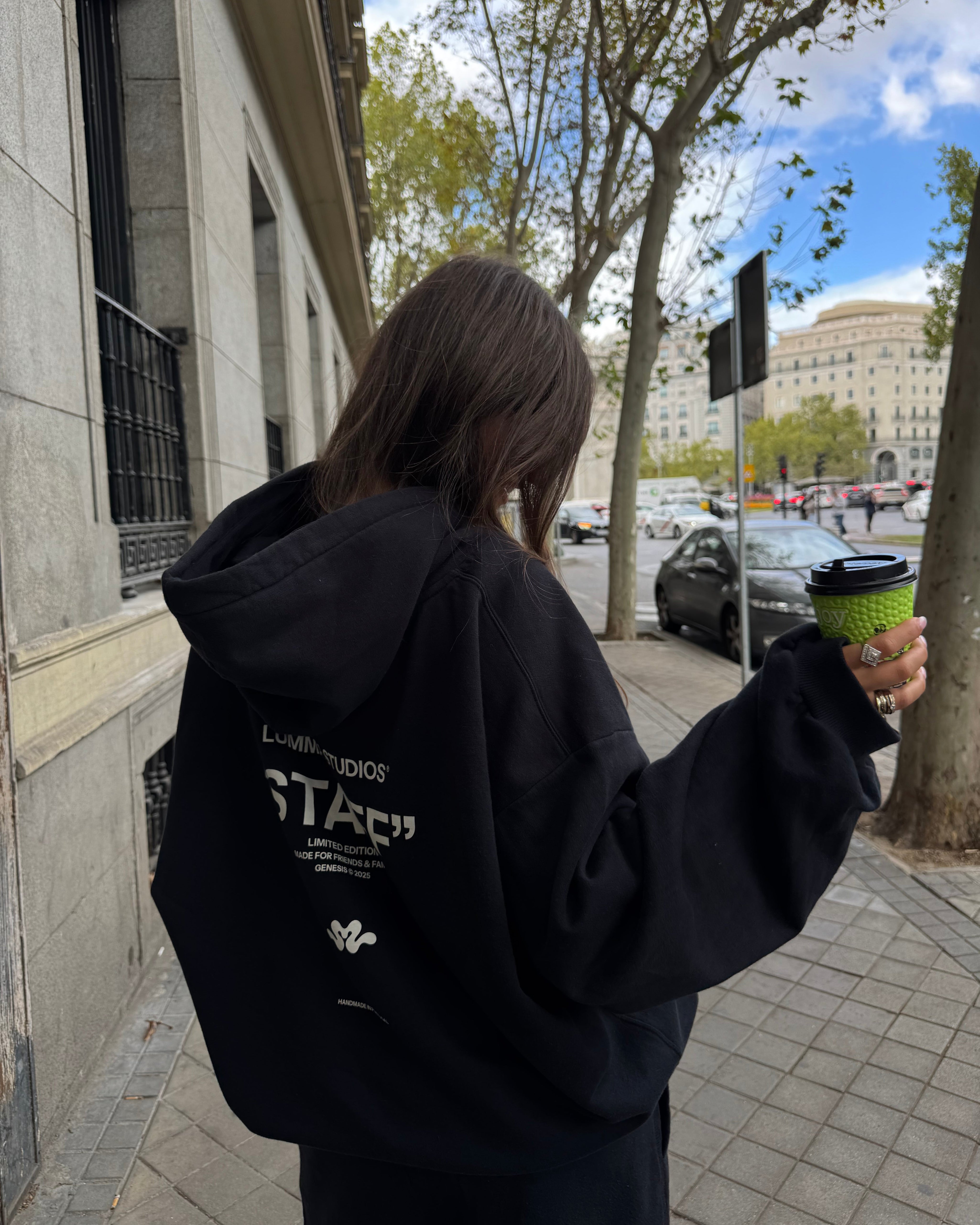Person wearing a black hoodie with text, holding a green cup outdoors.
