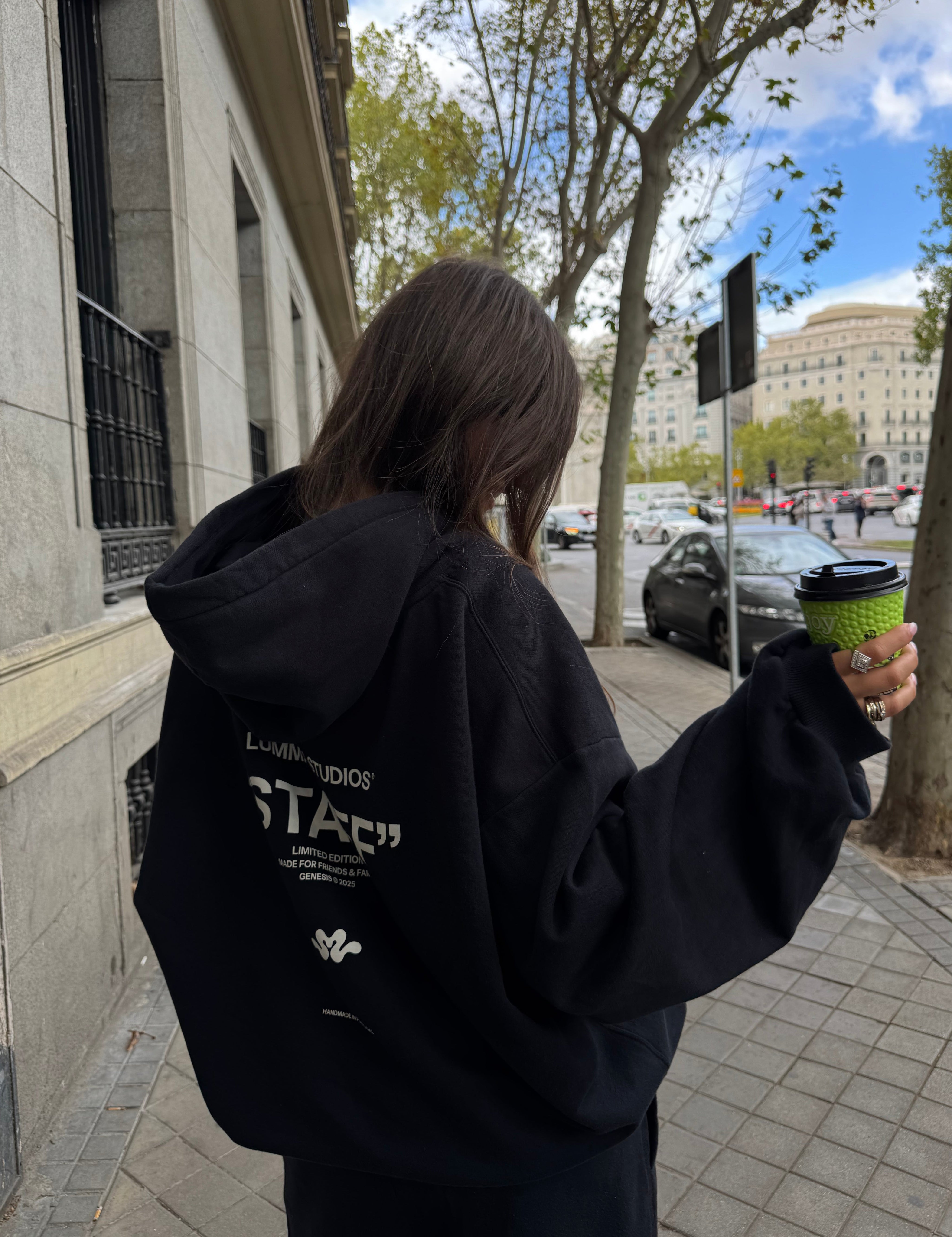 Person wearing a black hoodie with text, holding a green cup outdoors.