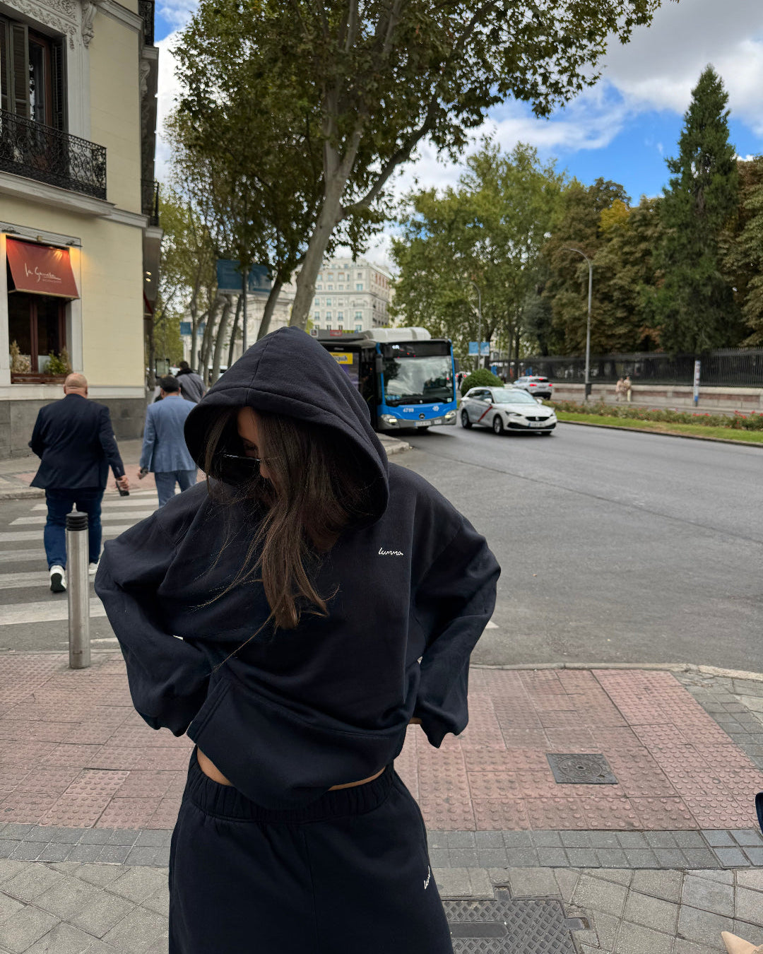 Person wearing a black hoodie on a street with trees and buildings in the background