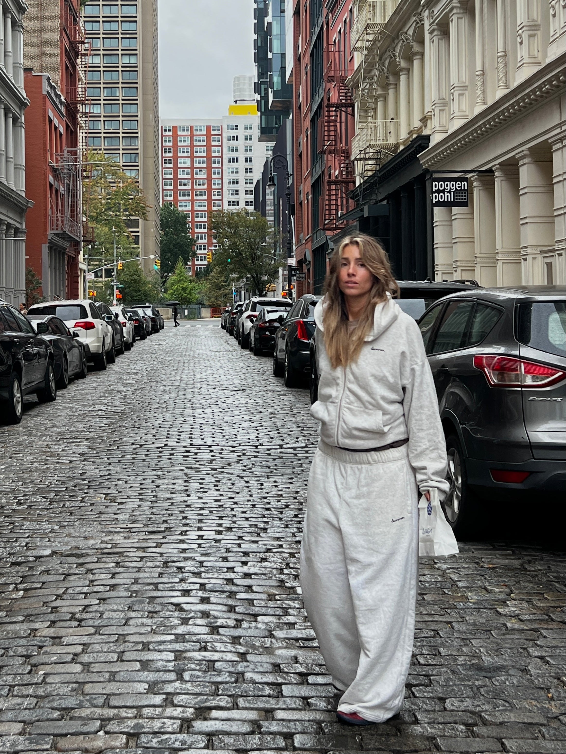 Woman walking down a city street with tall buildings and parked cars.